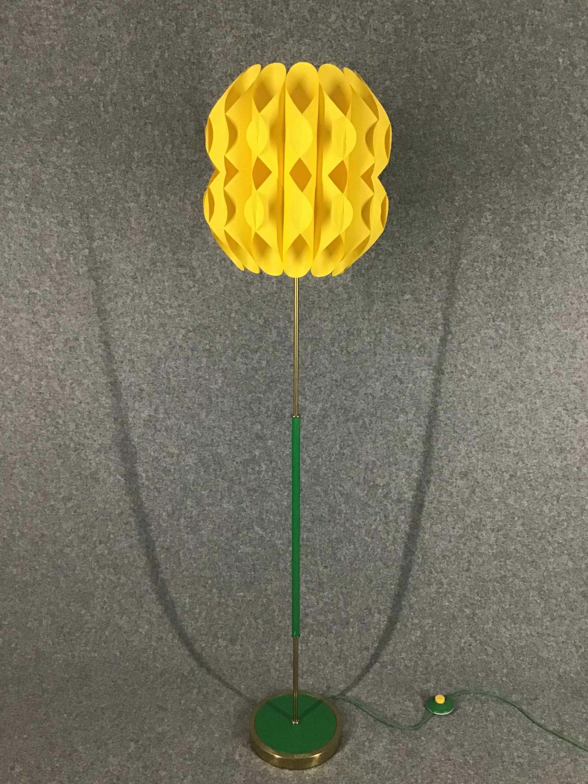 Stehleuchte mit einem leuchtend gelben Lampenschirm aus kunstvoll gefaltetem Material und einem schlanken, grünen Ständer auf stabilem Sockel